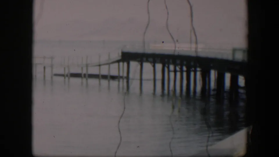 A 1951 Utah waterfront frame shows pier supports over calm water, a muted horizon, and pronounced vertical scratches from the 8mm source.