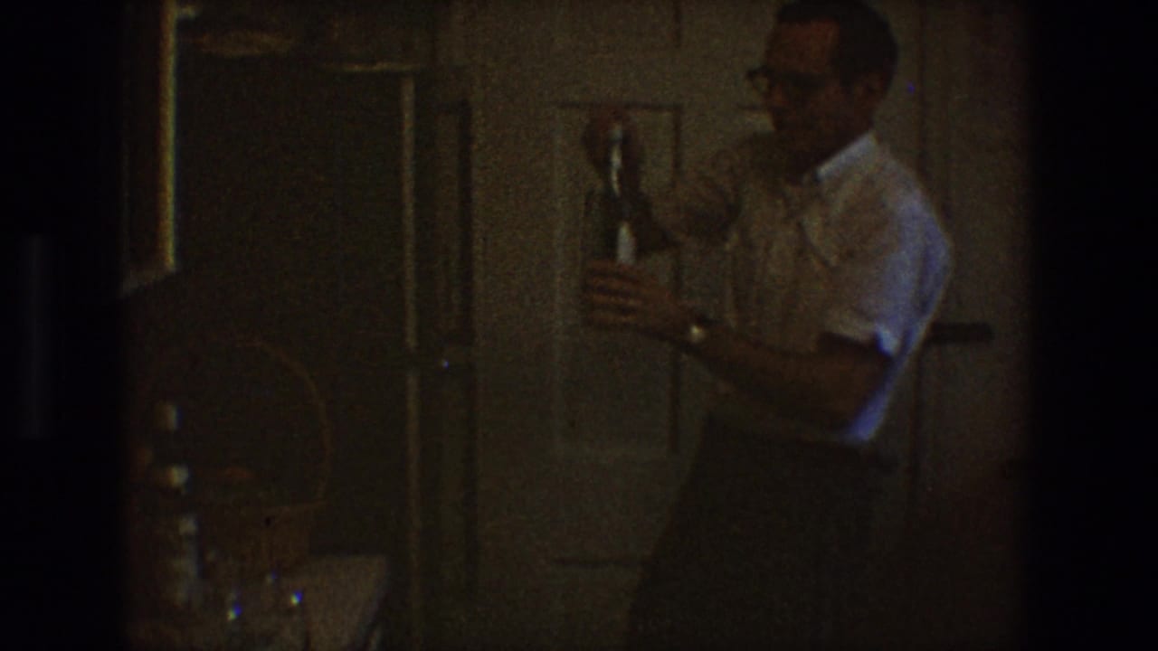 Man in a white shirt holds a bottle in a dark interior near paneled doors.