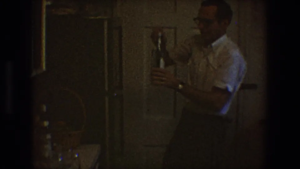 In a dim 1968 Toledo home-movie frame, a man in glasses and a white short-sleeved shirt holds a glass bottle indoors.