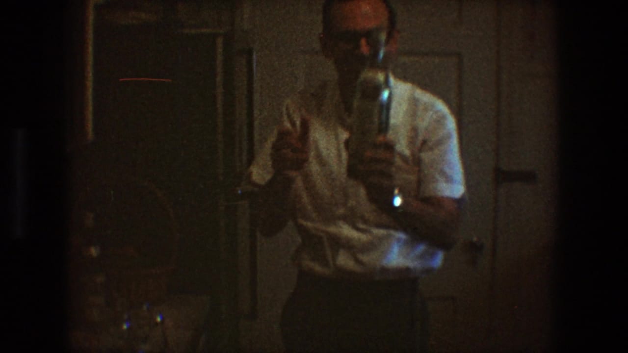 Man faces the camera while holding a bottle in a dim household room.