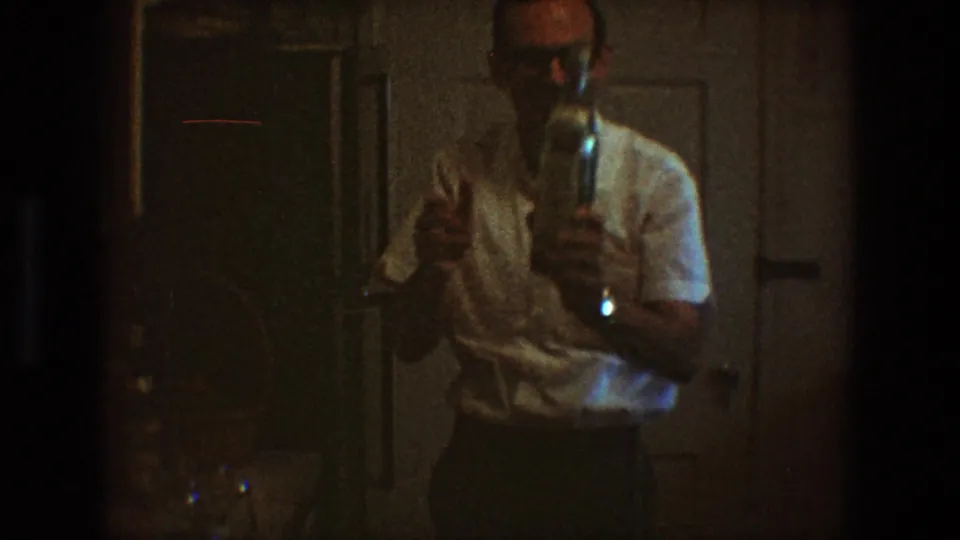 The man turns toward the camera in the dark interior, holding the bottle upright against paneled doors in 1968 Toledo footage.