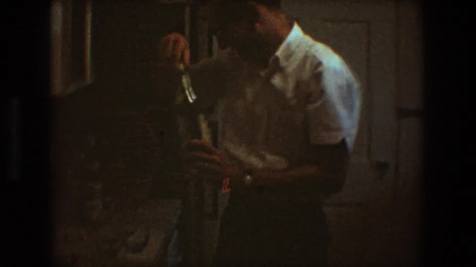 In the low-light 8mm frame, the man appears to examine or open the bottle while standing beside a table indoors.