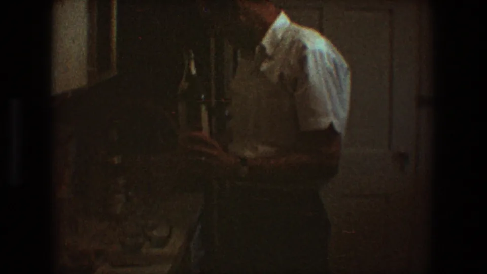 The man is seen in partial profile holding the bottle near paneled doors, with dark grain and warm color from the 8mm scan.