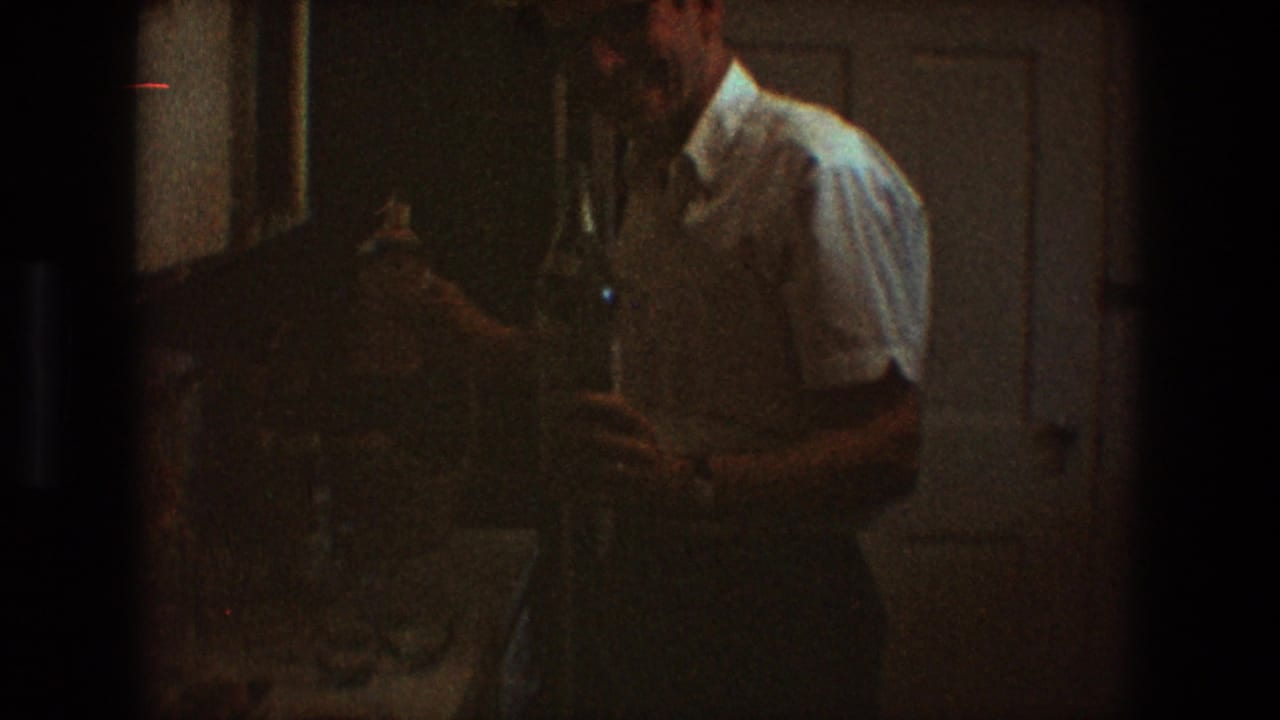 In a 1968 Toledo home interior, an adult man in a white short-sleeved shirt stands near kitchen cabinets with bottles in hand.