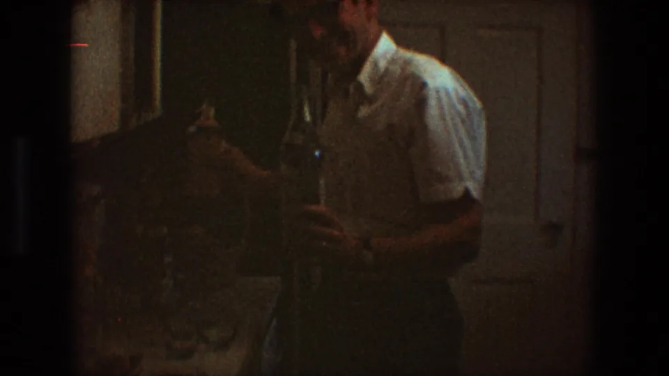 In a 1968 Toledo home interior, an adult man in a white short-sleeved shirt stands near kitchen cabinets with bottles in hand.