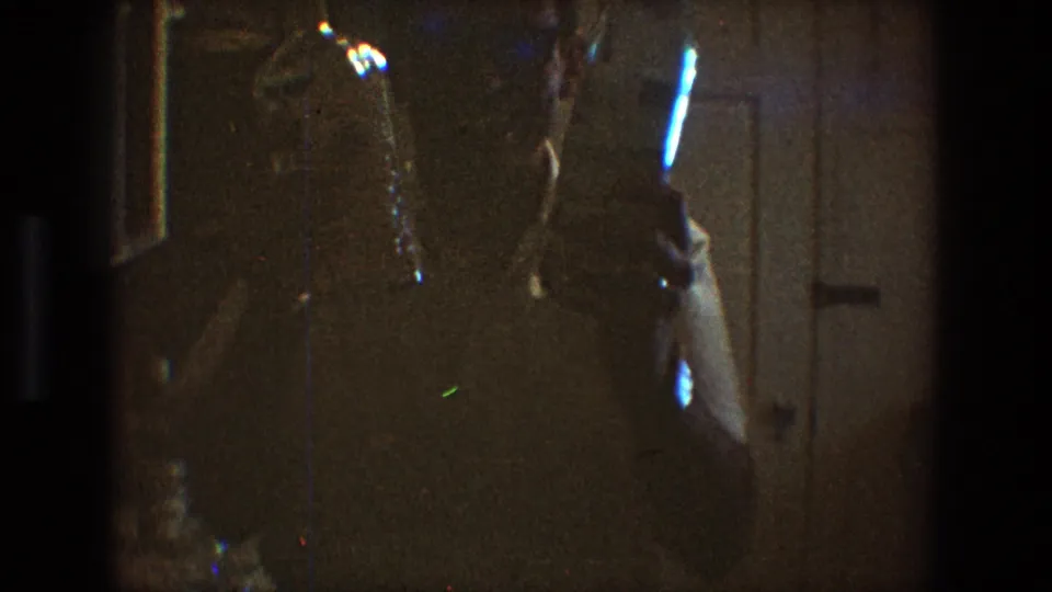 A low-light 8mm frame shows the man partly obscured as reflective bottle shapes catch light in the Toledo home interior.