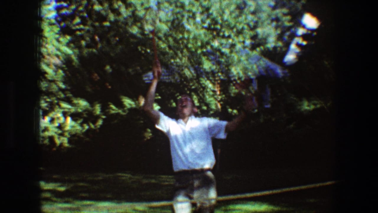 Man in a white shirt raises a badminton racket near a garden net.