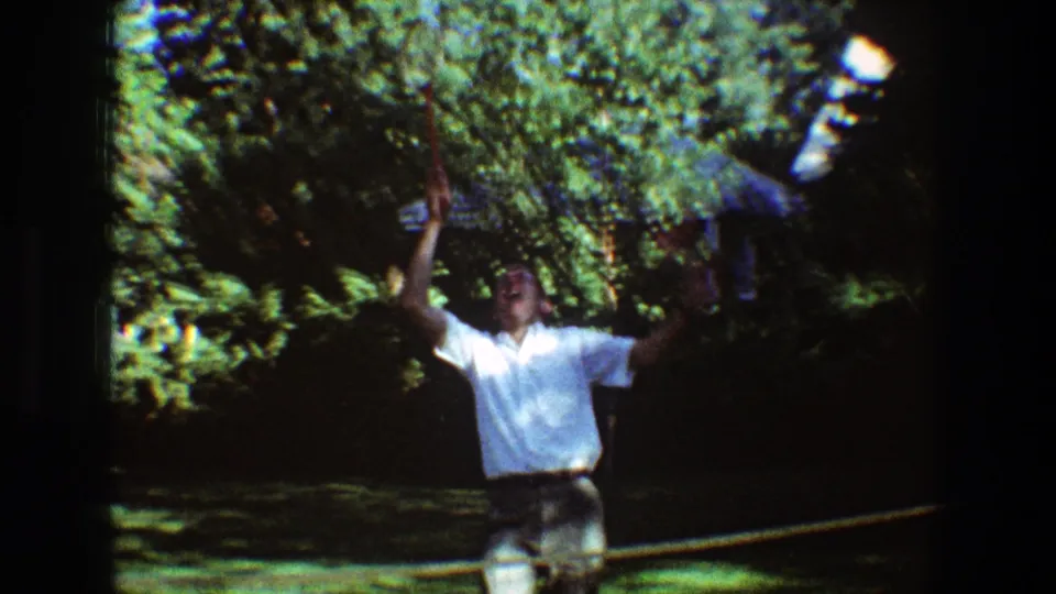 In 1968 Toledo home-movie footage, a man in a white shirt swings a badminton racket in a shaded garden.