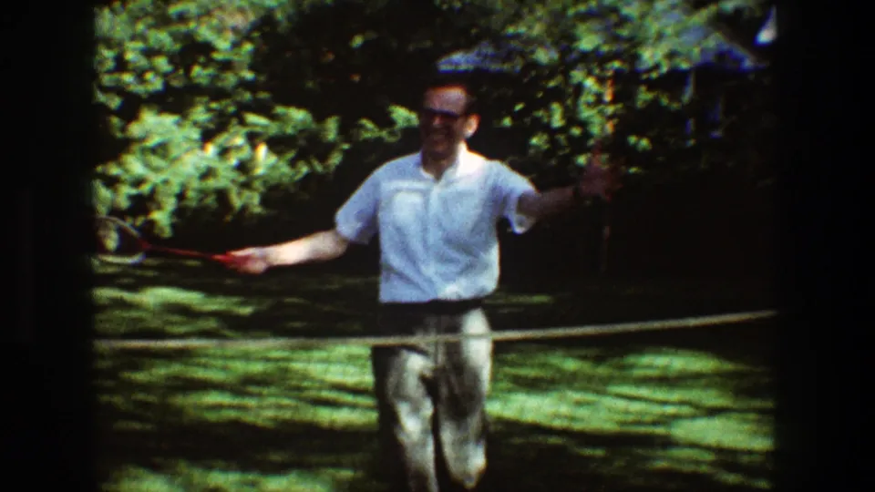 The badminton player lunges forward with his racket in a grainy 1968 garden home movie frame.