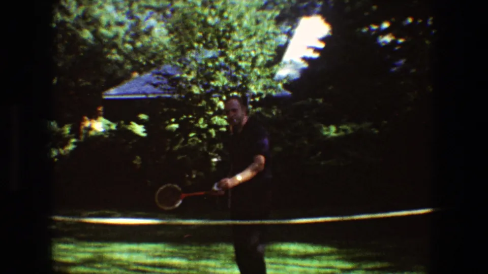 An older man in dark clothing holds a badminton racket on the opposite side of the garden net in Toledo.