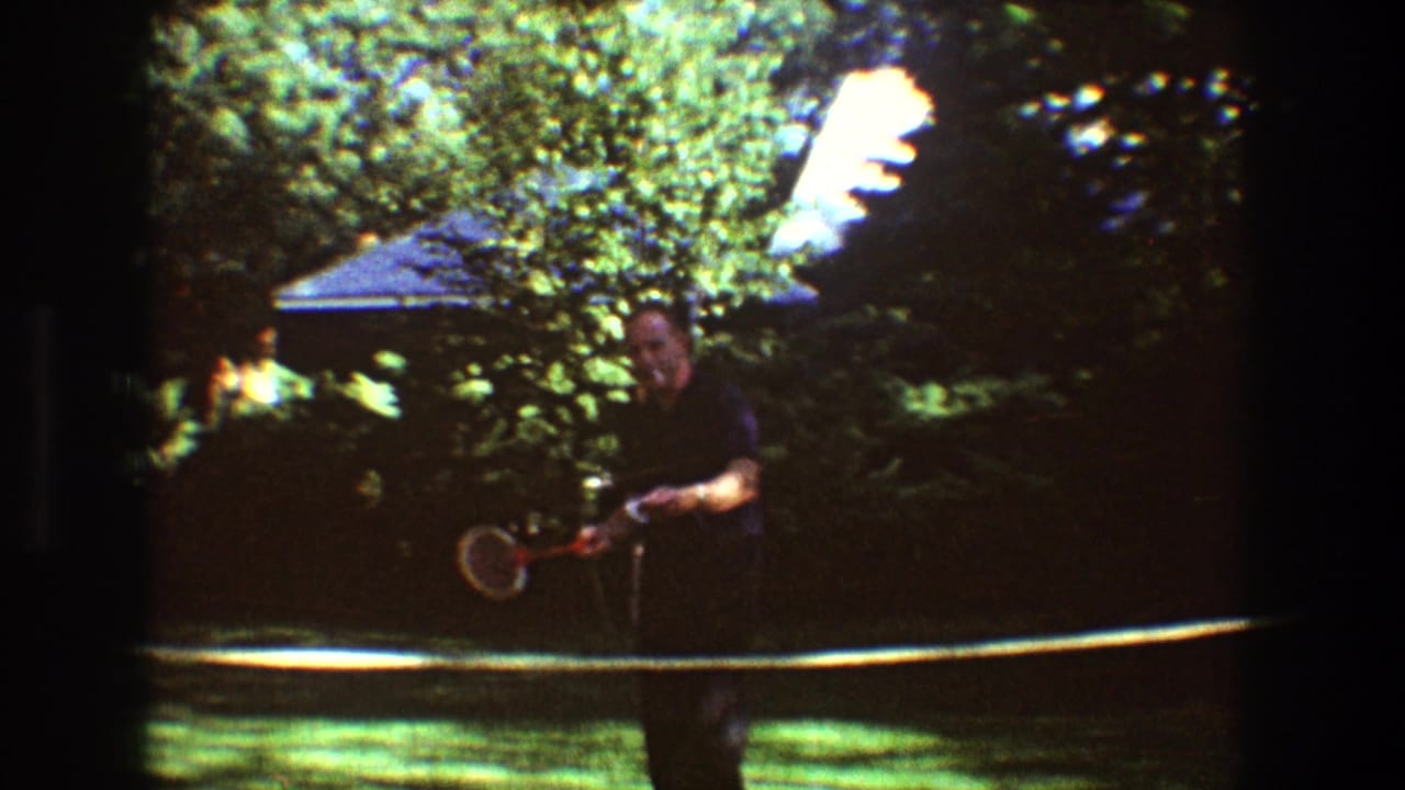 Man holding a badminton racket near a backyard net with trees and a house behind him.