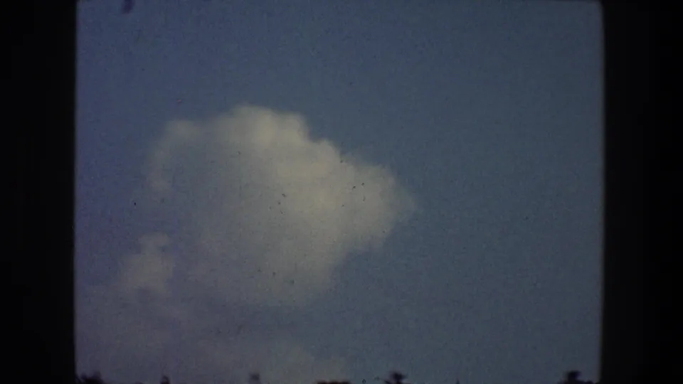 1968 Toledo 8mm frame showing a larger cloud mass in pale sky, with film grain, gate edges, and faint residential silhouettes at bottom.