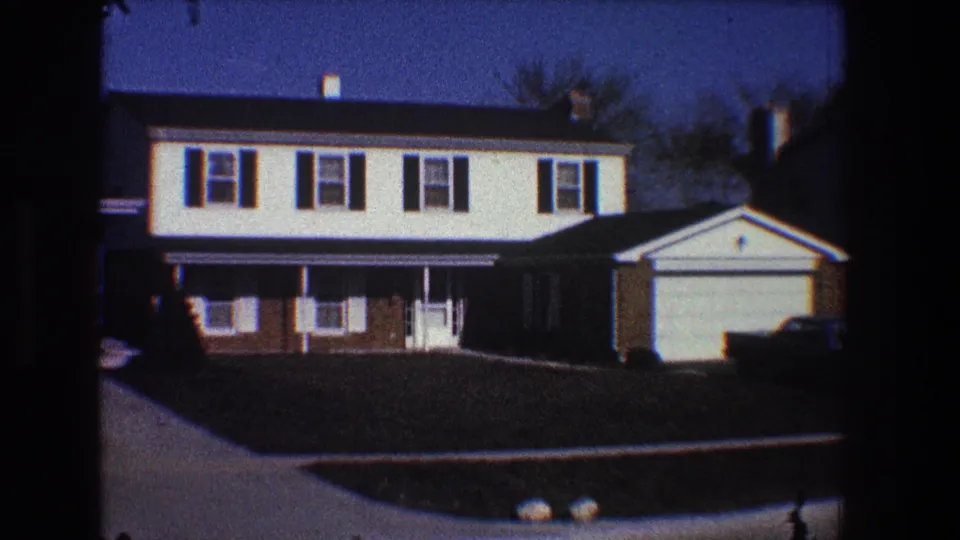 1968 Toledo frame of a suburban house and attached garage, with porch details, dark shutters, lawn, and a dark sedan partly visible near the driveway.