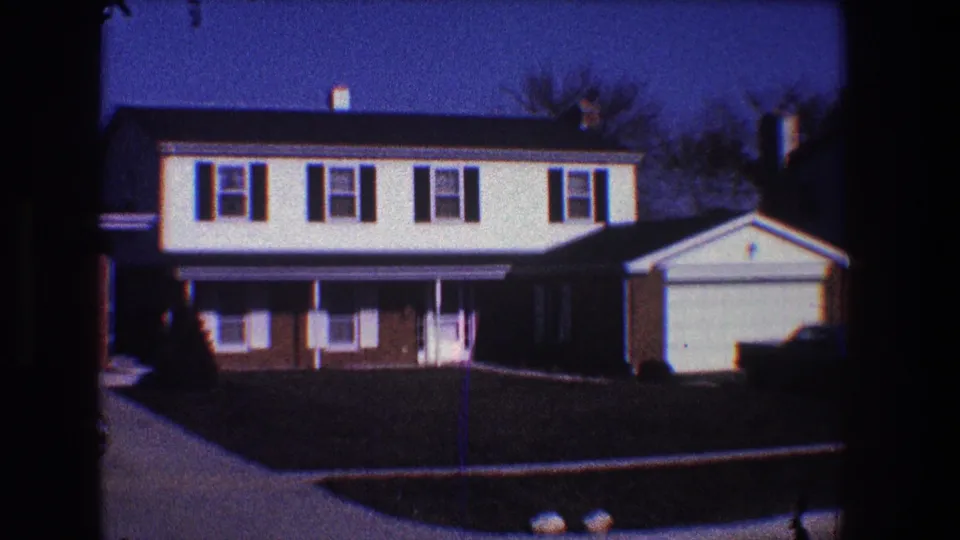 1968 Toledo 8mm frame showing the front of a suburban split-level house, attached garage, shaded lawn, porch columns, and blue sky.