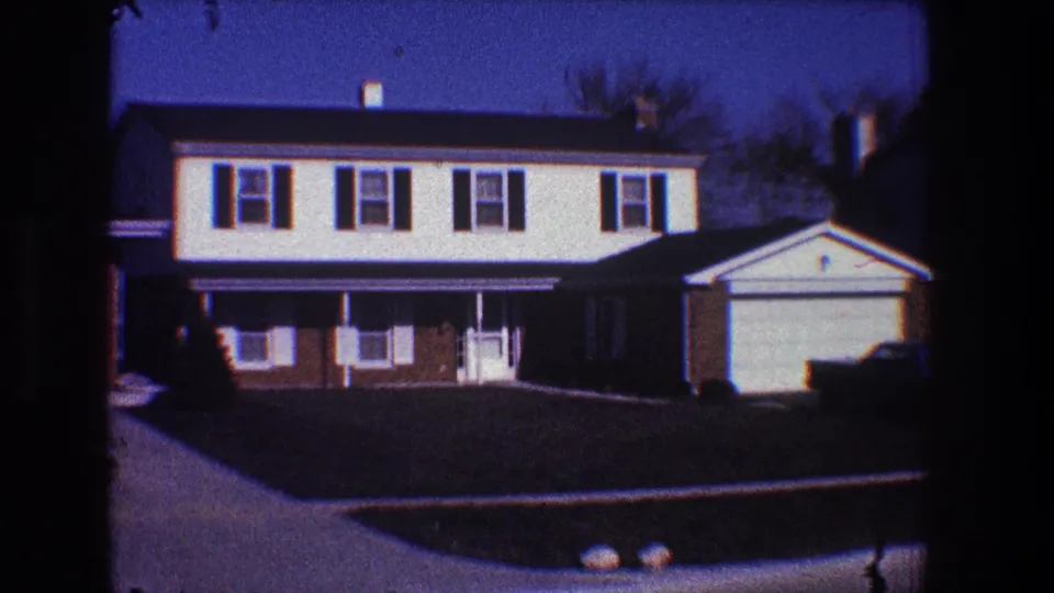 1968 Toledo home-movie frame of a suburban residence with dark shutters, attached garage, front lawn, driveway curve, and a parked dark sedan.