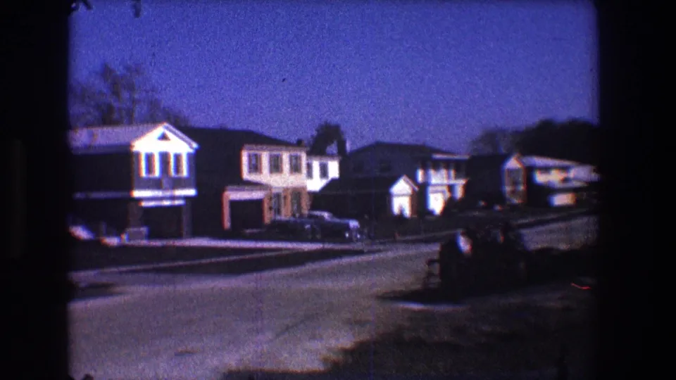 Late-1960s Toledo home-movie view of a suburban block with houses, garages, parked cars, lawns, and long shadows.