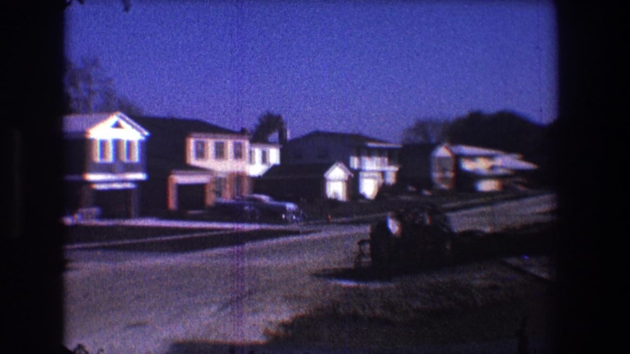 1968 Toledo home-movie frame showing a quiet suburban street with mid-century houses, driveways, and parked cars under a blue evening sky.