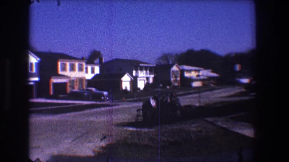 1968 Toledo frame of a quiet suburban block, with houses, garages, a parked car, lawns, and blue-tinted dusk light.