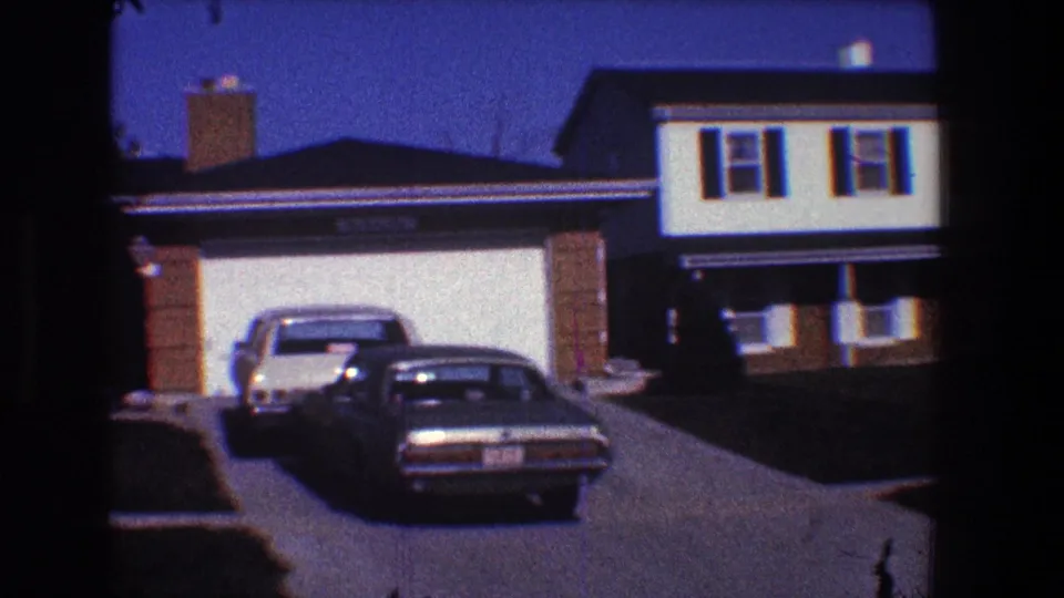 1968 Toledo suburban driveway with two parked cars, a garage, brick chimney, house facade, and neighboring home at dusk.