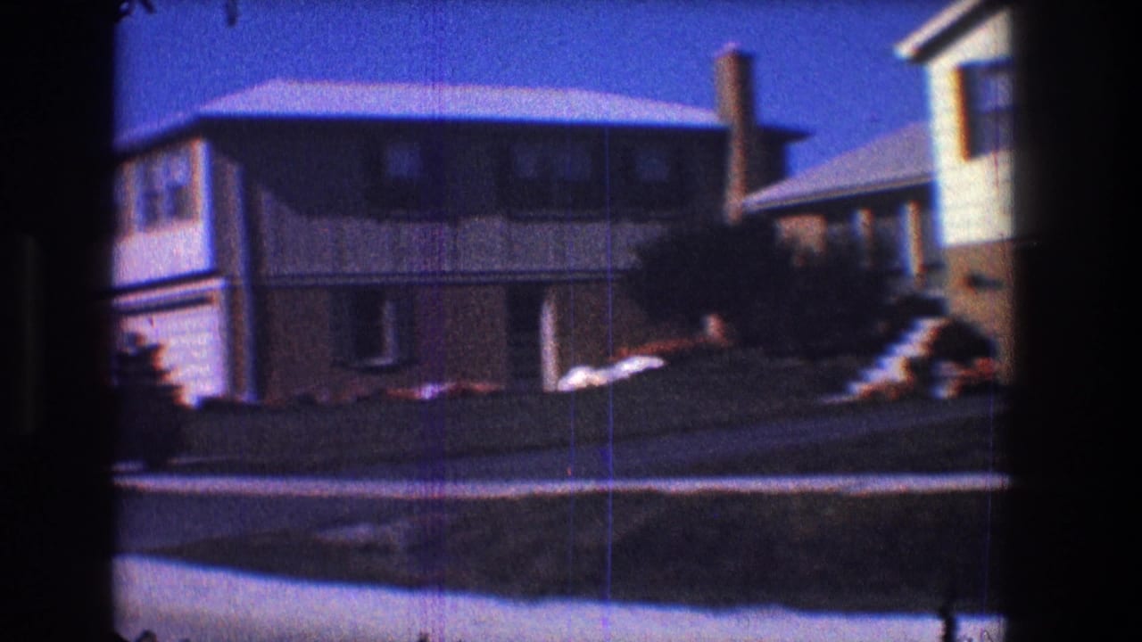 1968 Toledo home-movie frame showing mid-century suburban houses, lawns, and a quiet street edge under strong blue sky.