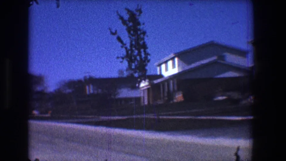 1968 Toledo frame with a low, slightly tilted view of a suburban house, lawn, street, and small tree in soft 8mm color.