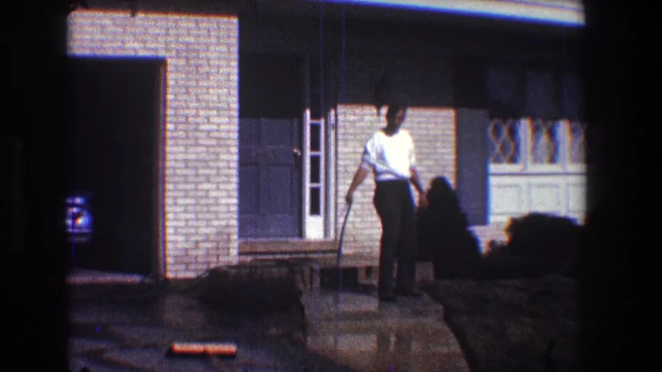 1968 Toledo home-movie frame showing a woman in a white shirt holding a leash beside a brick suburban house and dog.