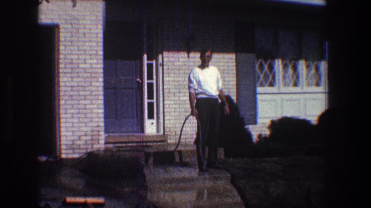 Man uses a hose on a wet walkway in front of a brick house.