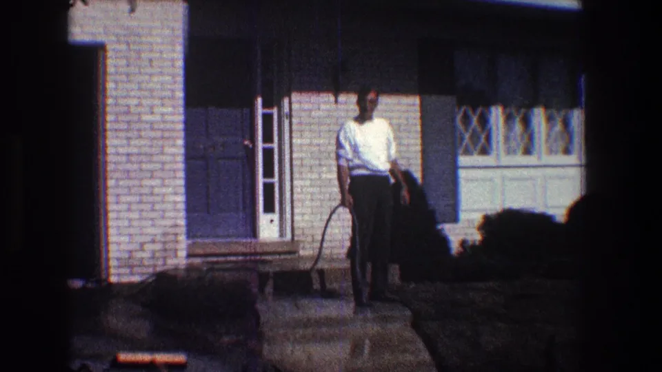 The adult man stands centered on the damp walkway with the garden hose in hand, framed by the brick house exterior, blue shutters, and bright 1960s home-movie sunlight.