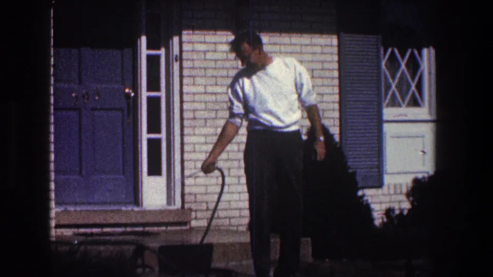 A closer 8mm frame shows the man looking down as he directs the garden hose near the concrete step, with the blue front door, shutters, brickwork, and window details behind him.