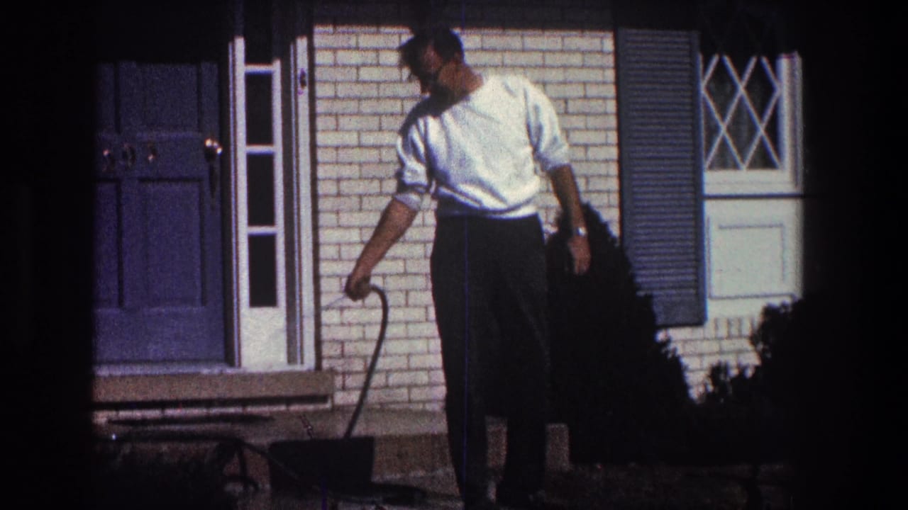 Man in a white shirt uses a hose wand near a brick porch and blue door.