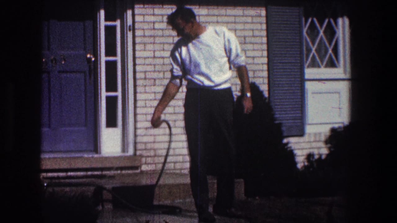 Man looks down while holding a small hose wand beside the brick house.