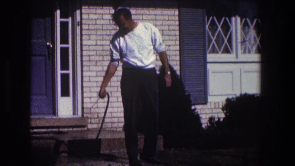 The closing frame keeps the man and hose wand centered against the brick exterior, blue door, and white-trimmed window.