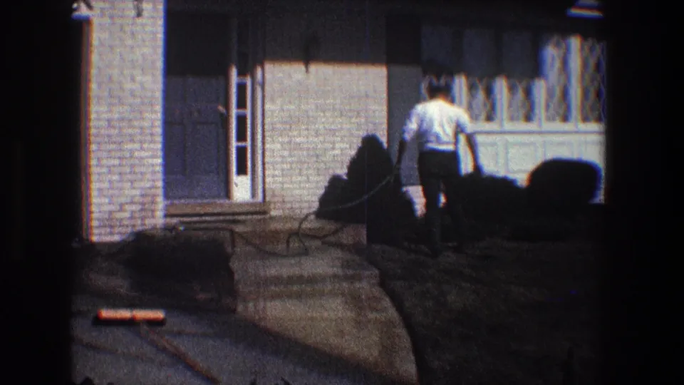 Seen from behind, the man cleans near the house entrance as the hose runs across the concrete walkway.
