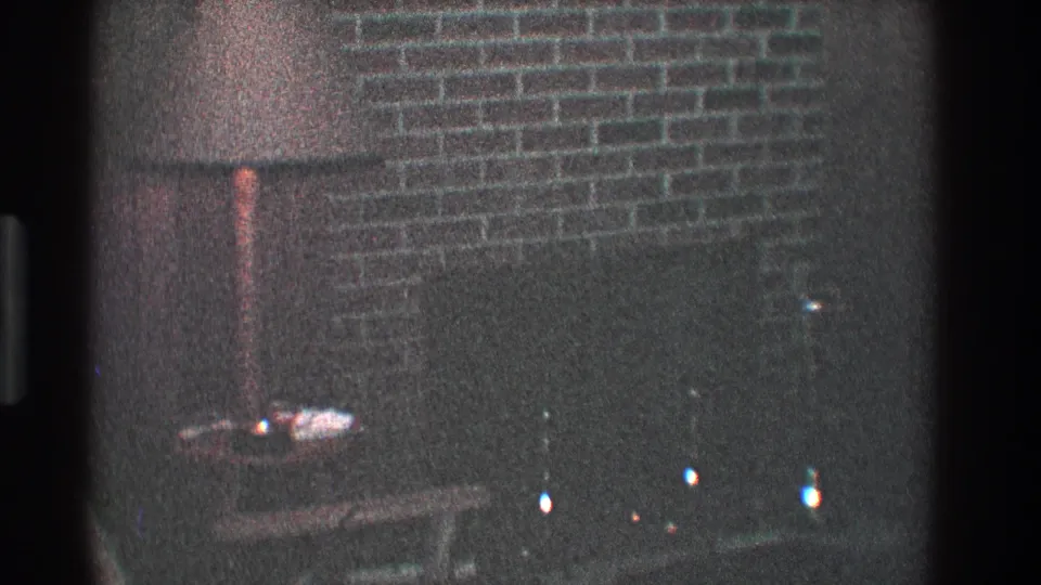 1968 Toledo home-movie frame showing a dim living room with a brick fireplace, floor lamp, small table, and dark hearth area.