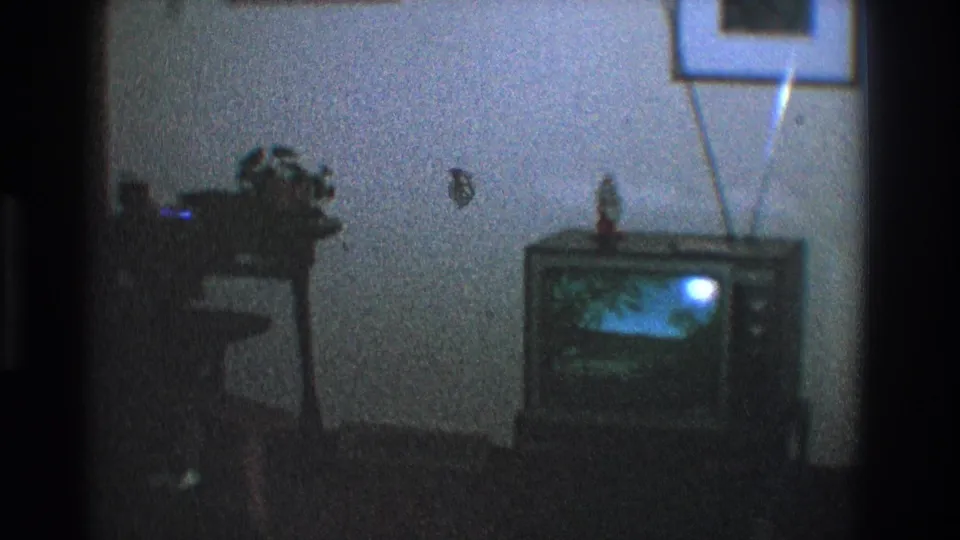 1968 Toledo home-movie frame showing a tube television with rabbit-ear antenna, a small object on top, and a nearby stand against a blue wall.