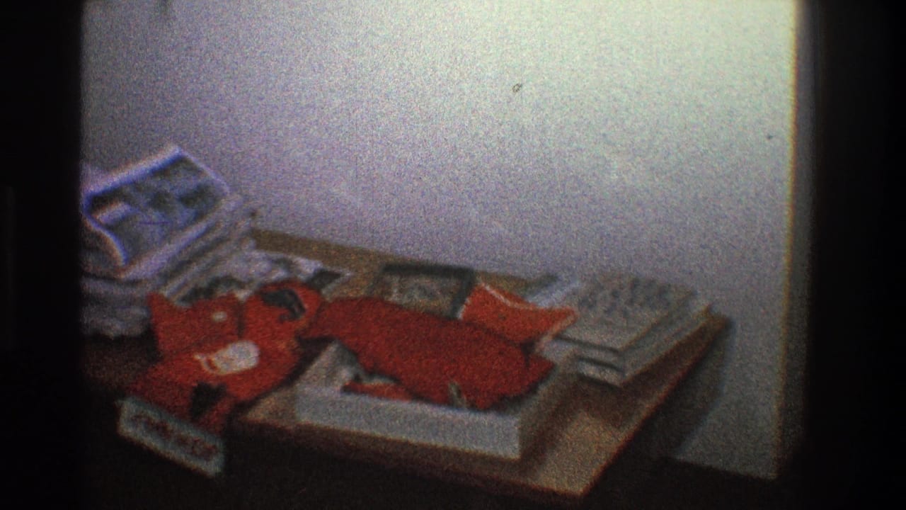 Cluttered wooden table with stacked papers and a red stuffed object in a dim room.