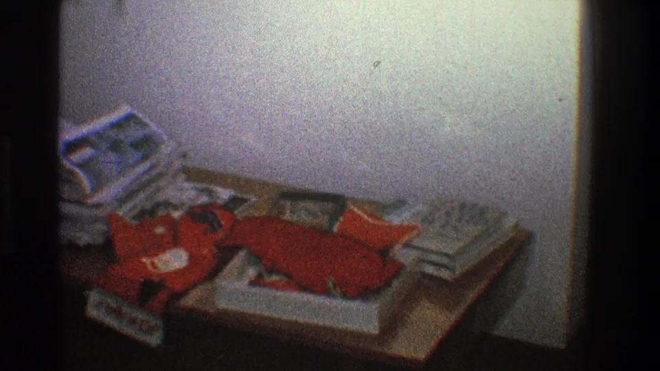 1968 Toledo home-movie frame showing a cluttered wooden table with papers, magazines, and a red stuffed object against a plain wall.