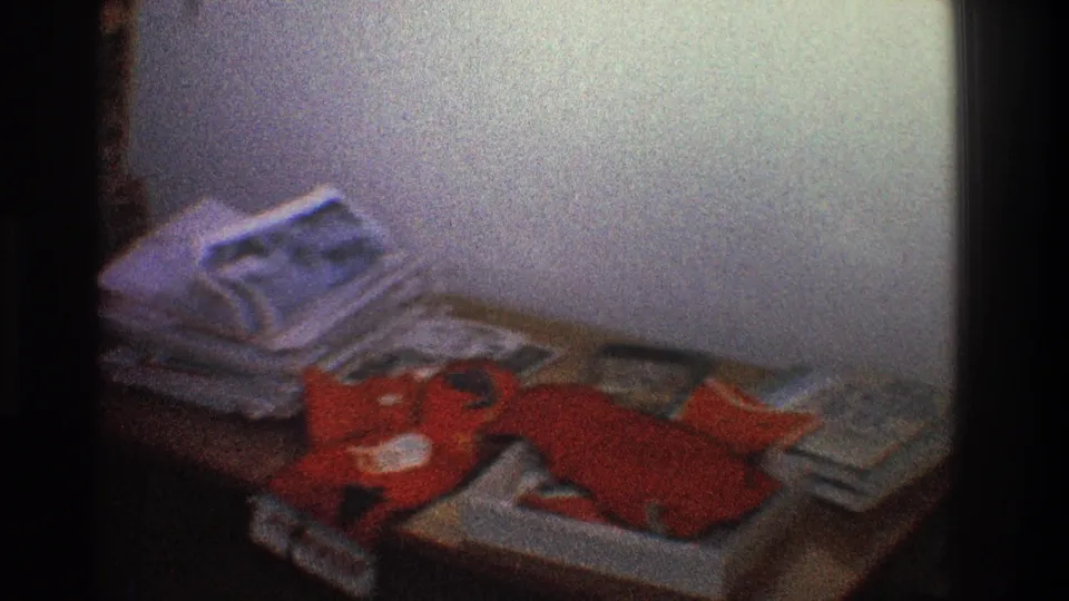 1968 Toledo interior frame with papers and magazines piled on a table beside a red stuffed object, filmed in soft 8mm light.