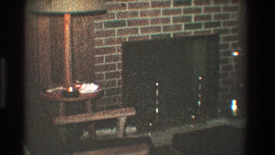 1968 Toledo home-movie frame of a brick fireplace with a dark hearth screen, small side table, and bench in low interior light.
