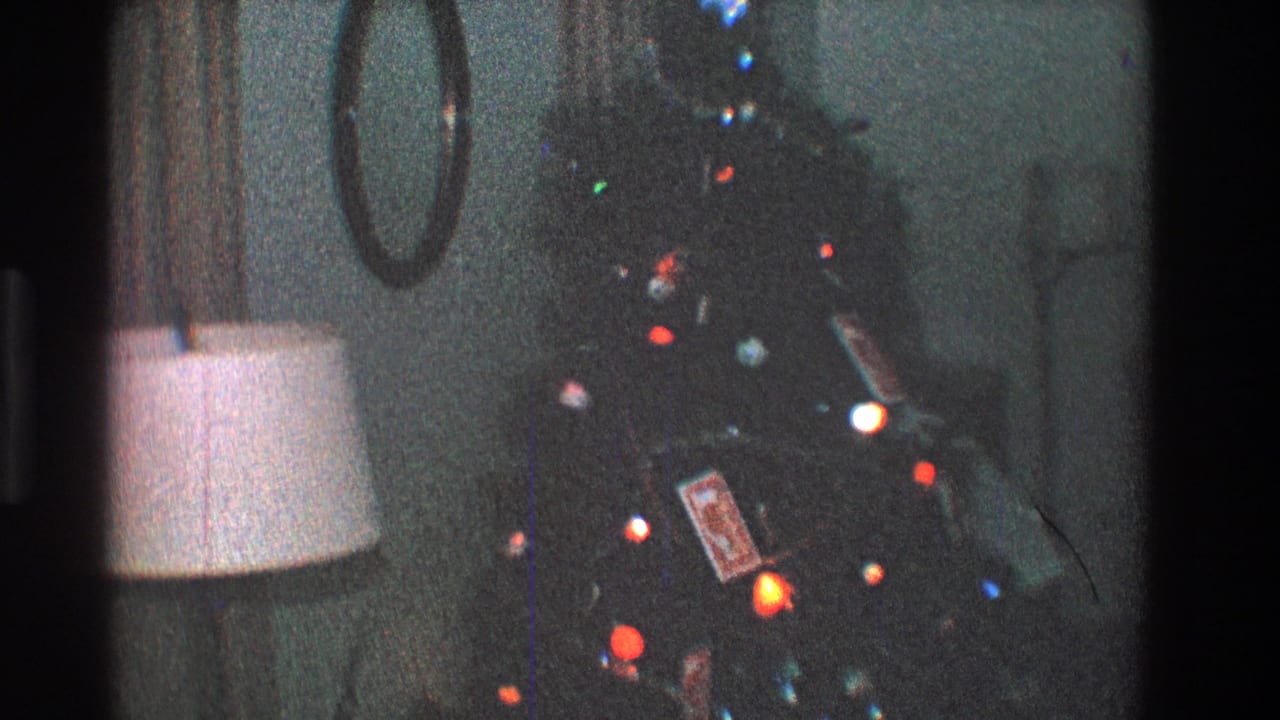 Dim living room with a decorated Christmas tree, colored lights, lamp, and wall mirror.