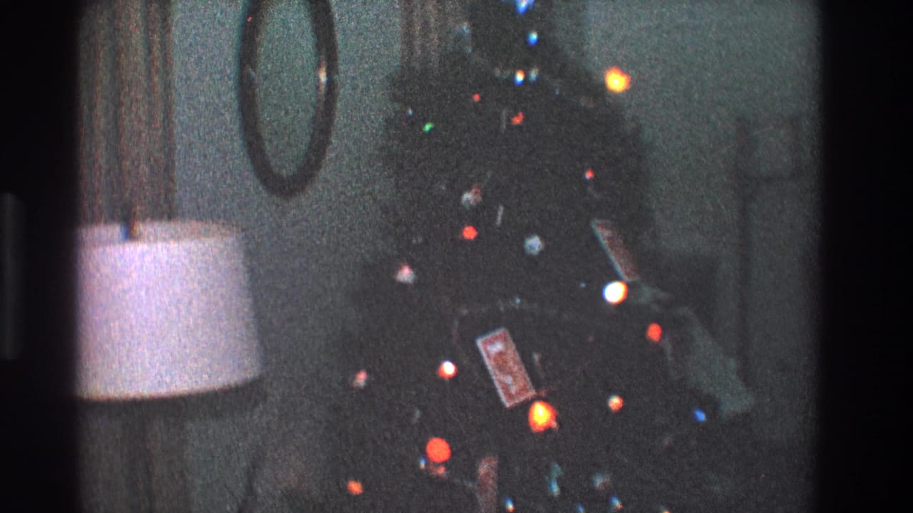 1968 Toledo home-movie frame showing a dim living room Christmas tree with colored lights beside a white-shaded lamp and round wall mirror.