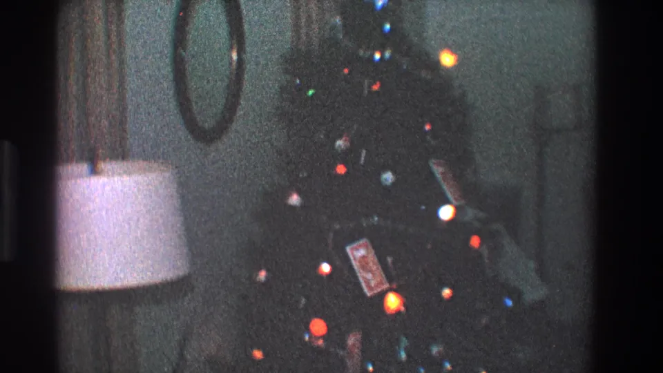 1968 Toledo home-movie frame showing a dim living room Christmas tree with colored lights beside a white-shaded lamp and round wall mirror.