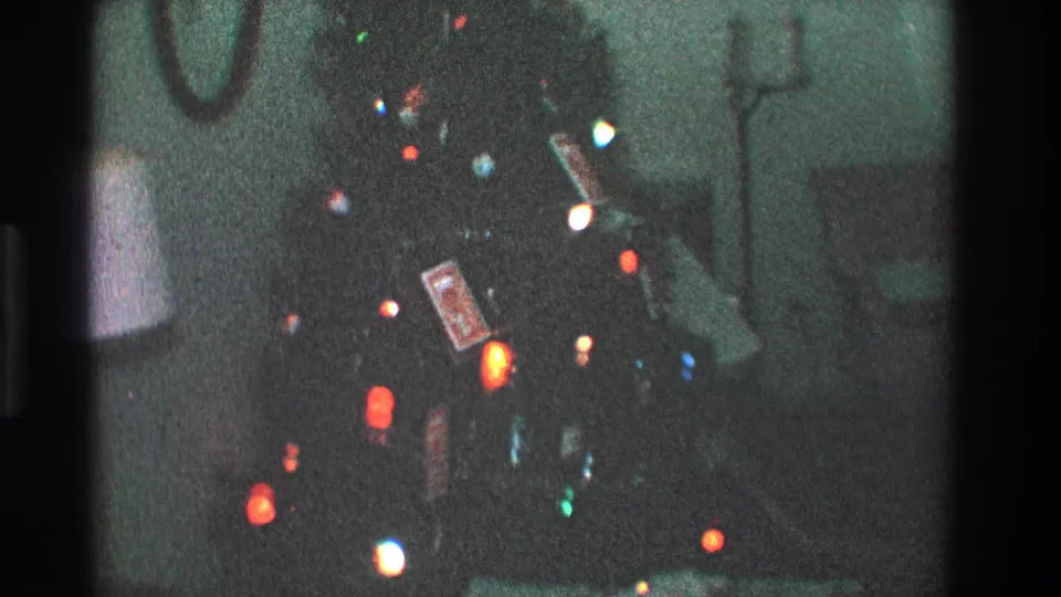 1968 Toledo home-movie still showing colorful Christmas tree lights and ornaments in a softly exposed living room with a lamp and wall mirror.