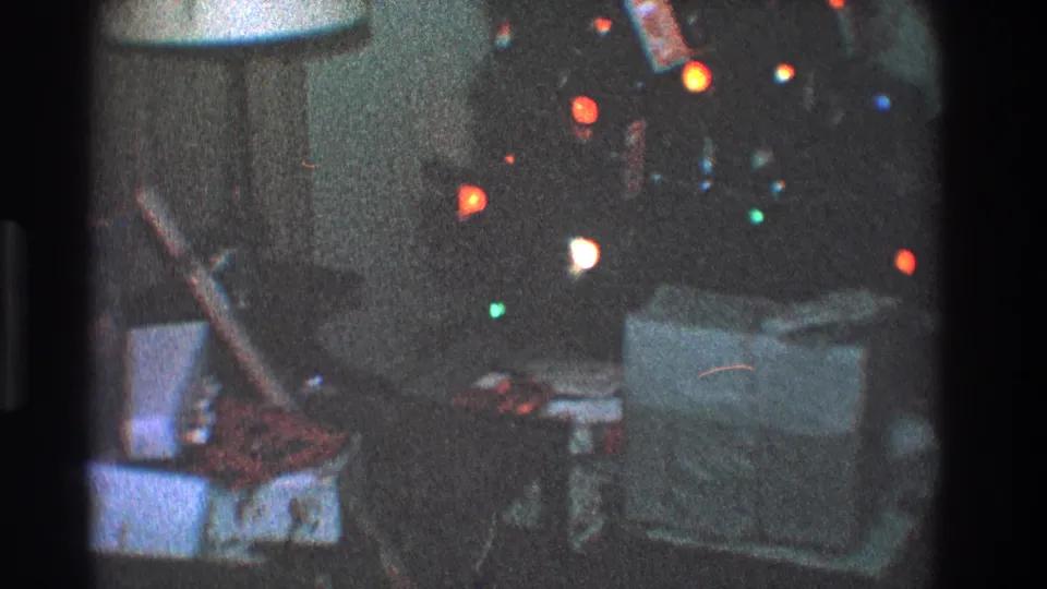 1968 Toledo home-movie frame showing wrapped gifts on a couch near colored Christmas tree lights and a white-shaded lamp.
