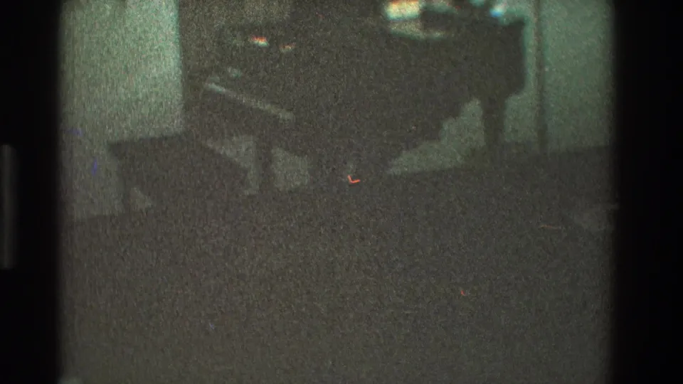 1968 Toledo home-movie frame shifting toward dark living-room furniture, possibly a piano with a bench or chair.