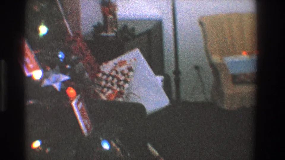 Grainy 1968 Toledo frame showing Christmas tree lights, a wrapped present, lamp, and pale orange chair in a home interior.