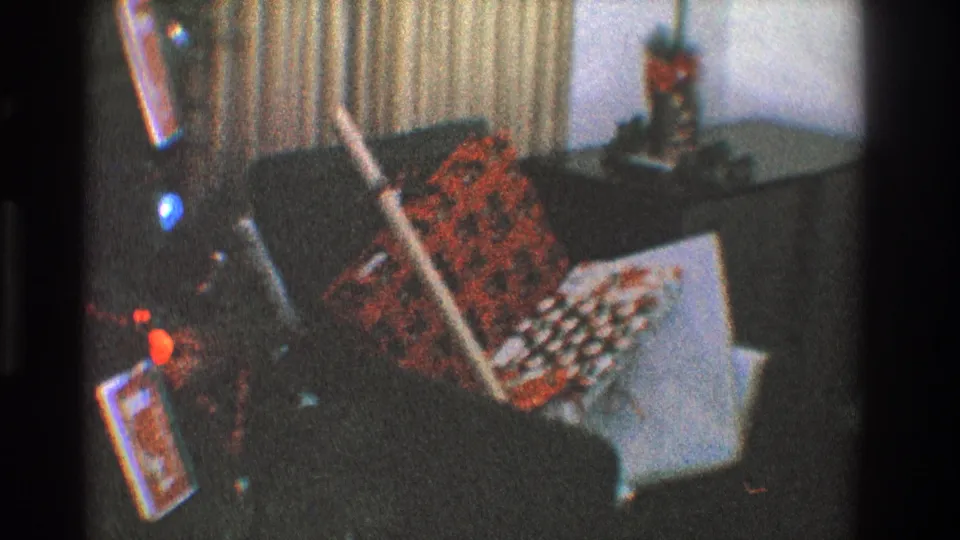 1968 Toledo home-movie frame with a tree edge, gift packages, a white box, and patterned holiday textile beside dark furniture.