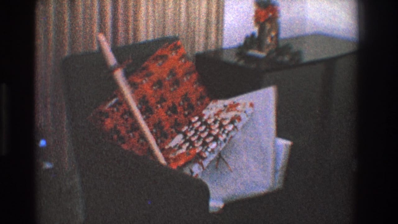 Dark armchair with patterned pillows and a broom leaning against it near striped curtains.