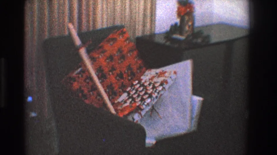 1968 Toledo home-movie frame showing a dark lounge chair with red and white patterned textiles and a broom leaning against it.