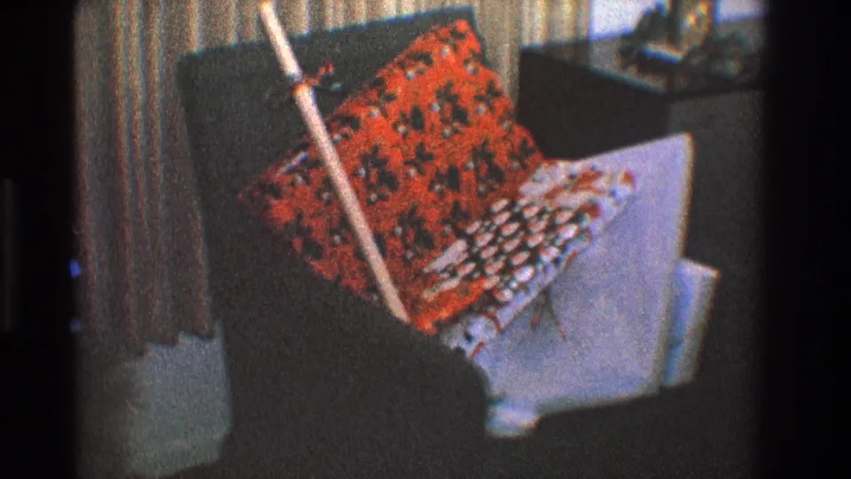 1968 Toledo interior frame with a dark armchair, red patterned cushion, white textile, leaning broom, and side table in the background.